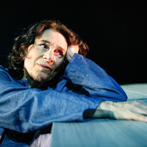 Gisela Nilsson porträtterar sin mamma i föreställningen Hitta Hem på Teater Västernorrland. Kvinna lutar sig mot scenkant av gammalt trä och blickar hoppfullt bortåt.