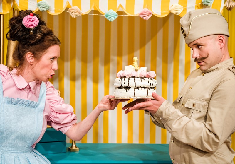 Kvinna i kitschig kostym och man i militäruniform och mustasch presenterar en maräng tårta med ljus.