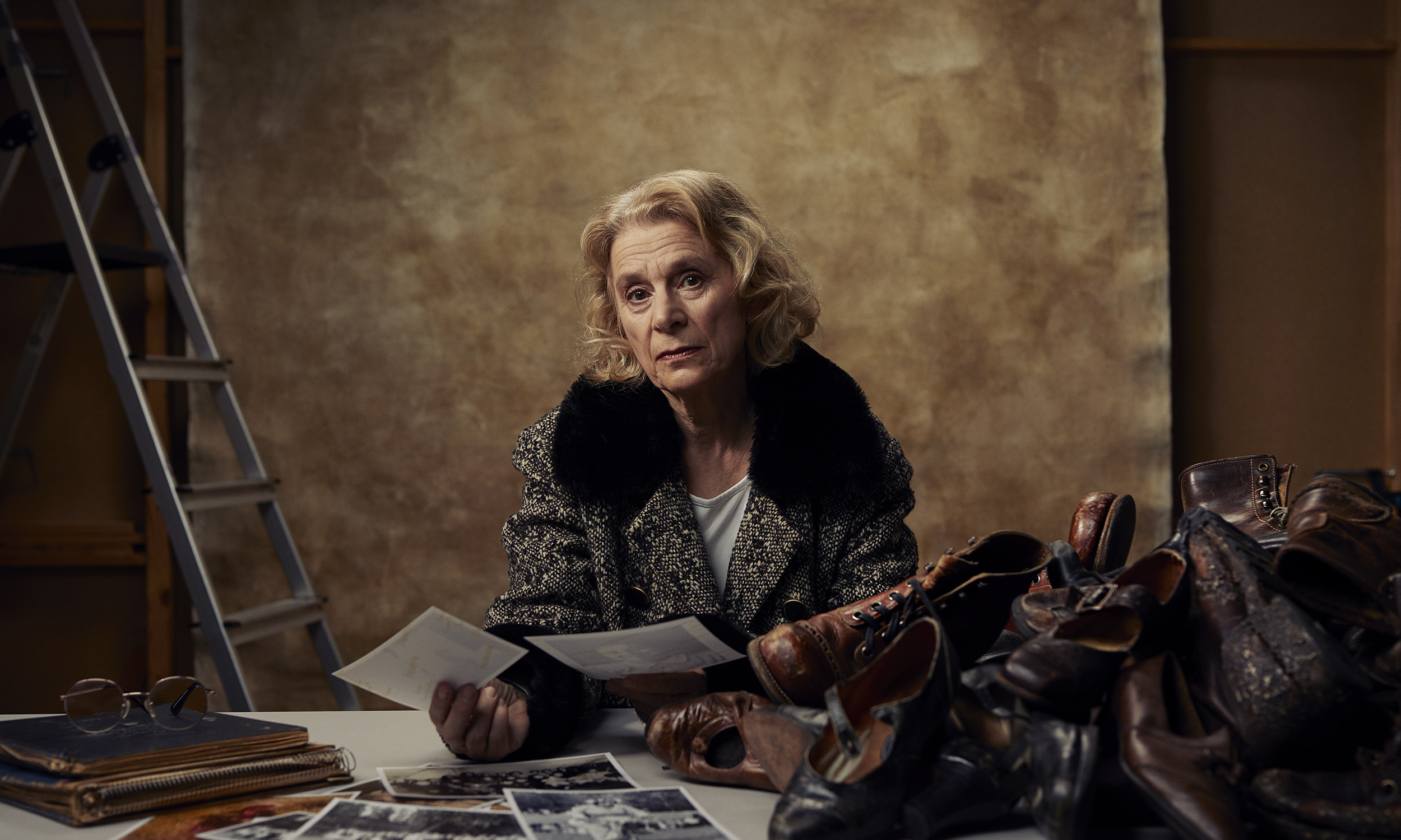 Affisch bild till Teater Västernorrlands föreställning Hitta Hem. Huvudrollsinnehavaren Gisale Nilsson sitter vid ett bort med en hög judiska skor och sin mammas gamla brev och dokument.